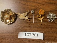 Five assorted brooches laid on wooden surface including flower with pearl, bird, small rose pin, gold rose, and silver flower cluster.