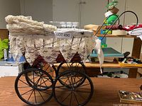 Photo of decorative wicker baby carriage with lace canopy, showing side view with four large metal spoked wheels and deep basket.