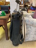 Photo showing green golf bag with multiple compartments and attached carrying strap, containing golf clubs with metal shafts and various club heads including drivers and irons.