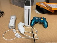 Nintendo Wii console, white Wii remote controller with silicone sleeve, white Nunchuk controller, and blue GameCube controller placed on a table.
