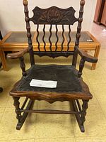 Front view of a vintage wooden rocking chair with black leather seat and carved backrest showing floral detail. The leather seat is visibly worn and not sturdy.