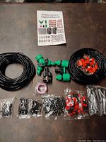 Complete view of the kit showing black tubing coils, green hose connectors and splitter pieces, plastic fittings, drip emitters, plumbing tape, plastic stakes, and instruction manual.