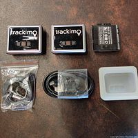 Two Trackimo GPS tracking units with magnetic backs, two USB charging cables in plastic bags, and one white silicone protective case on a brown surface.