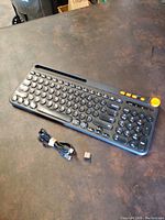 Full view of keyboard with round black keys, orange function keys, USB charging cable, and small Bluetooth dongle placed beside it.