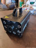 Front angle showing black metal fog machine with handle, front LED lights, and fog nozzle.