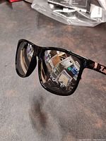 Glossy black framed sunglasses with silver mirrored lenses and chrome accents on arms, taken on dark surface.