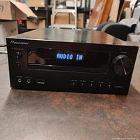 Front view of Pioneer X-HM21V compact amplifier powered on displaying 'AUDIO IN', showing volume knob, power button, USB port, headphone jack, and control buttons.