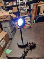 Full view of the black LED lamp with a round head on adjustable stand, plugged in and turned on on a table in a storage room.