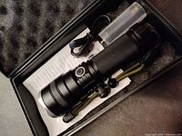 Flashlight in open box showing USB charging cable, battery holder tube, and instruction manual inside protective foam case.