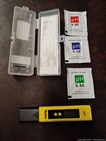Yellow digital water pH meter tester and pH buffer powder packets with a transparent plastic storage case on a brown surface.