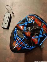 Front view of the black zombie mask showing blue EL wire stitching and red blood-like paint. Battery pack with on/off switch visible.