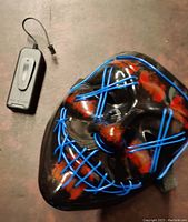 Front view of the black plastic zombie mask with blue LED wiring and red blood-like paint, showing battery pack with on/off switch.