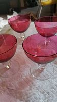 Four cranberry colored stemmed glasses on a white cloth, showing their bowl and stem detailing, suitable for drinks or desserts.