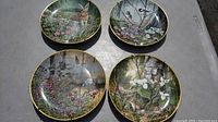 Four decorative plates displayed on a flat surface showing nature scenes including animals and flowers