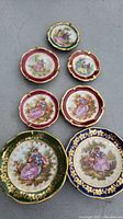 Full view of all 7 mini Limoges plates arranged on a surface with visible romantic couple designs and rich gilt borders