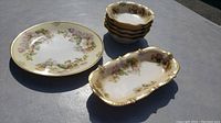 Seven pieces of antique Prussian dishware shown with floral designs and gold trim on porcelain.