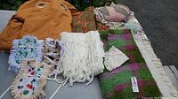 Wide view of orange curtain panel, three bottle aprons, Cushendale throw, knit scarf, doily, and quilt style throw pillow laid out