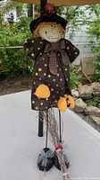 Front view of the Halloween witch decoration standing on a white surface outside, showing details of the fabric dress, pumpkin decoration, hat, and broom.