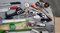 Top-down view of various kitchen utensils including knives, whisks, pizza cutters, skewers, measuring cups, wooden spoons, baster, and peelers laid out on a surface.