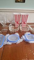 Clear plastic storage containers with blue seal lids arranged on wooden surface, two red and white striped plastic popcorn containers visible in background, four clear plastic drinking glasses behind containers.