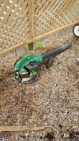 Full view of gas-powered handheld Weed Eater leaf blower placed on dry ground with wood lattice fence in background.