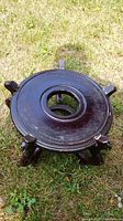 Top and angled view of round dark wood plant stand placed on grass, showing circular recessed center and four legs.