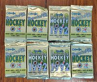 Photo showing eight unopened 1992 O-Pee-Chee Premier hockey card packs, arranged in two rows of four. Packs are metallic with prominent text indicating the brand, year, and number of cards per pack.