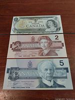 Three Canadian banknotes: 1-dollar bill from 1973 on top, 2-dollar bill from 1986 in middle, 5-dollar bill from 1986 on bottom. Each bill displays front side with portraits and denomination.