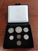 Photo shows open black coin case with seven coins displayed on velvet lining and logo on lid.