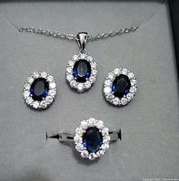 Photo showing a jewelry set consisting of a silver-tone necklace with an oval blue stone pendant bordered by white stones, matching oval blue stone earrings bordered by white stones, and a matching ring featuring the same stone pattern.