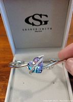 Silver toned bracelet with a rainbow-colored heart-shaped centerpiece and small white stones, displayed inside a George Smith branded jewelry box.