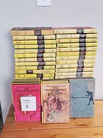 Photo shows 56 Nancy Drew books stacked in three piles with mostly yellow spines featuring a circular portrait illustration. In front are three books with colorful illustrated fronts visible: one pink cover titled 'The Crooked Banister', a beige cover titled 'The Strange Message in the Parchment', and one blue cloth hardcover titled 'The Password to Larkspur Lane'.