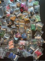 Photo shows a large number of small plastic bags laid out on a dark surface containing buttons in various colours and styles, including pink, green, blue, red, and natural tones.