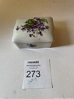 Top and side view of white porcelain trinket box decorated with purple flowers and green leaves, with auction lot number card.