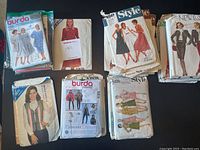 Photo showing a variety of vintage women's sewing patterns laid out on a wooden surface, including Burda, Style, New Look, Kwik Sew, and Simplicity brands. Envelopes show illustrations of women's clothing styles from mid to late 20th century, featuring dresses, skirts, blouses, and jackets.