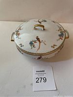 Top, side view of the round covered serving dish showing the bird and floral painted design and gold handle on the lid.