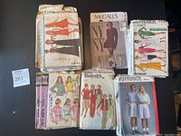 Photo showing multiple vintage McCall and Butterick sewing patterns with illustrated women in various garments such as dresses and pants. The patterns show wear and some are stacked.