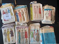 Photo of vintage Simplicity patterns arranged on a dark surface, showing various pattern envelopes with illustrated women's fashions, some envelopes worn and torn.