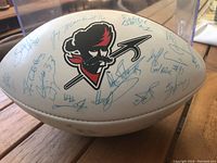 View of one side of the white Wilson football showing Ottawa Rough Riders pirate logo and multiple blue ink signatures including players with numbers like #85