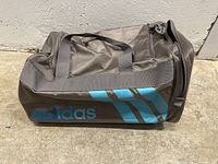 Photo showing side view of gray Adidas gym bag with blue Adidas logo and stripes, nylon fabric with two handles.