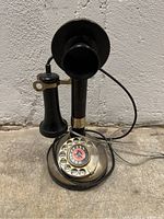 Front view of antique candlestick telephone showing rotary dial with numbered and lettered holes and a black bird emblem on a red background in the center.