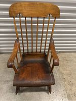Front view of antique wooden rocking chair showing curved top backrest, spindles, armrests and seat with visible wear.