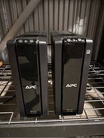 Two APC Back-UPS XS 1300 black uninterruptible power supplies standing side by side on metal shelving.