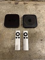 Two black Apple TV units with two aluminum Apple TV remotes on a dirty concrete floor.