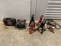 Wider view of all items together: four car jacks, battery jump starter, and cables on concrete floor.