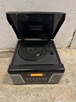 Black Crosley device with open lid revealing a turntable and tone arm for vinyl records, resting on a smooth floor surface.