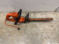 Full side view of orange electric hedge trimmer showing handle, blade, and power cord.