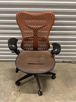 Front view of brown Herman Miller office chair showing perforated mesh backrest, mesh seat (damaged), black padded armrests, and black five-wheeled base.