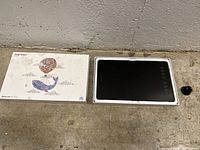 Photo showing the Huion drawing tablet alongside the original packaging box featuring colorful whale artwork, and the pen holder on a concrete floor.