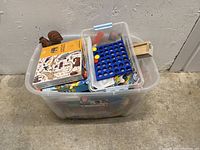 Large transparent plastic bin filled with assorted kids toys and games, viewed from above and side angle, showing a blue Connect Four game and various books and toys inside.